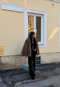 Manteau en fausse fourrure marron, haut noir court et pantalon noir à jambes larges ; tenant un sac à main matelassé bleu sarcelle. Décor urbain avec un mur beige en arrière-plan.