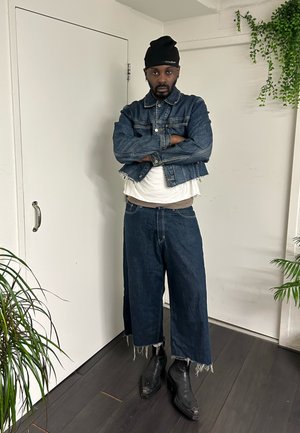 Hombre con gorro negro, chaqueta vaquera corta, camisa blanca, vaqueros anchos con bajos deshilachados y botas negras, de pie en el interior con los brazos cruzados.