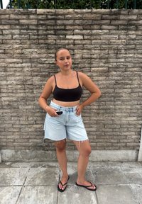 Musta lyhyt toppi, vaalean siniset farkkushortsit repsottavilla helmalla ja mustat varvassandaalit. Seisoo teksturoidun kiviseinän edessä.