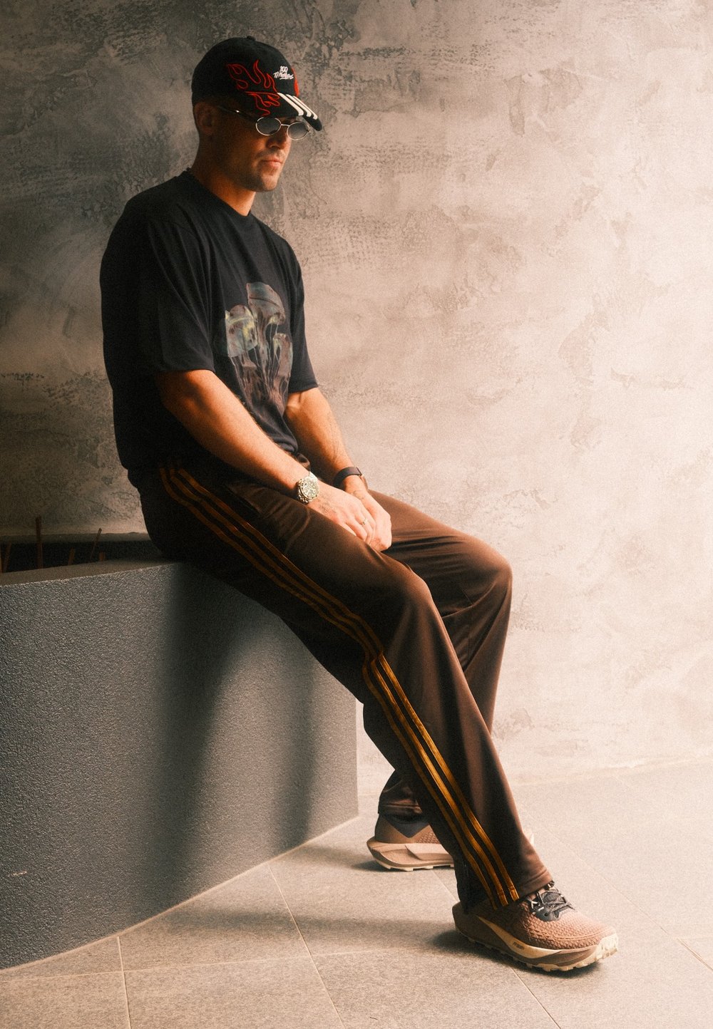 A man dressed in a black graphic t-shirt, brown track pants featuring orange stripes, sneakers, sunglasses, and a black cap is seated on a grey ledge.
