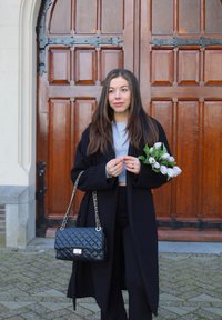 Juoda siuvinėta rankinė su sidabriniu grandinės dirželiu; moteris laiko baltas tulpes; dėvi ilgą juodą paltą ir kasdienę pilką palaidinę; medinės durys fone.