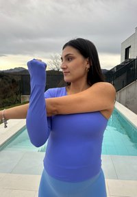 Γυναίκα με μπλε μακρυμάνικο τοπ που τεντώνει το δεξί της χέρι μπροστά στο στήθος δίπλα σε πισίνα σε εξωτερικό χώρο, με βουνά στο βάθος.