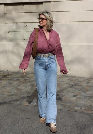 Mujer rubia con gafas de sol, blusa rosa, vaqueros azul claro, llevando un bolso marrón tejido, caminando por una calle empedrada a plena luz del día.