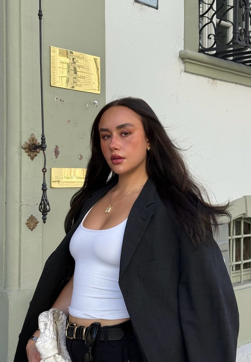 Veste noire oversize portée sur un débardeur blanc, avec un pantalon noir taille haute. Les accessoires comprennent un collier en or et un sac blanc texturé.