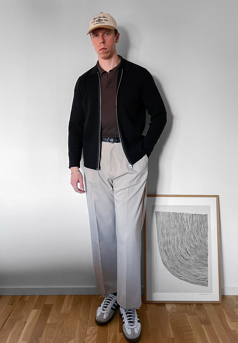 Man in beige pet, zwarte jas, bruin shirt, lichtbeige broek en sneakers staat naast ingelijste zwart-witte abstracte lijntekening op houten vloer.