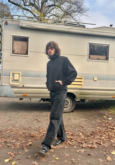 Chaqueta negra con cremallera, jeans negros holgados y zapatos negros brillantes. El fondo presenta una furgoneta camper gris claro con ventanas cerradas. Hojas de otoño en el suelo.