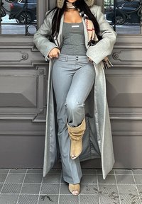 Personne en manteau gris, haut gris et pantalon gris levant une jambe pour montrer des bottines ouvertes en daim beige, debout devant une porte décorative grise.