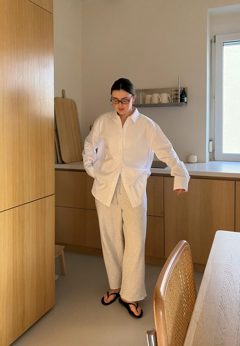 Vrouw met een bril, wit overhemd, grijze joggingbroek en zwarte sandalen, staand in een houten keuken vlak bij een raam en een eettafel.