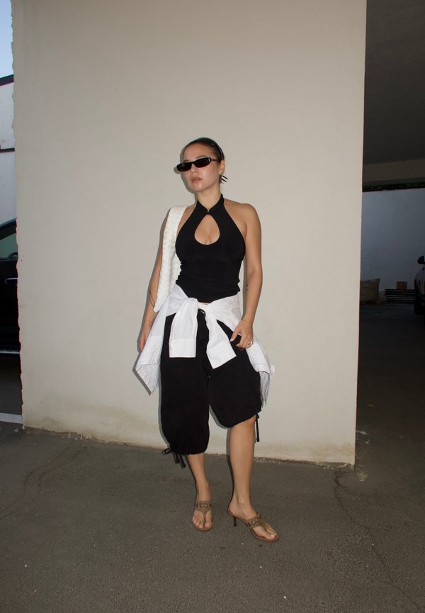 Femme en haut dos nu noir et pantalon à jambes larges avec une veste blanche nouée à la taille, portant des lunettes de soleil et des sandales à talons, debout dehors.
