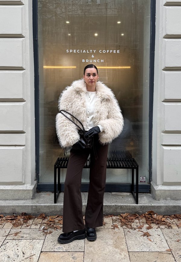 Übergroßer cremefarbener Kunstpelzmantel, weißer pullover mit Knöpfen, braune Nadelstreifenhose, schwarze Plateauschuhe, hält eine schwarze Handtasche.