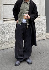 Cappotto lungo nero sopra una camicia a bottoni verde oliva, t-shirt bianca e jeans neri larghi. Scarpe da ginnastica grigie. Tiene in mano una tazza di caffè.