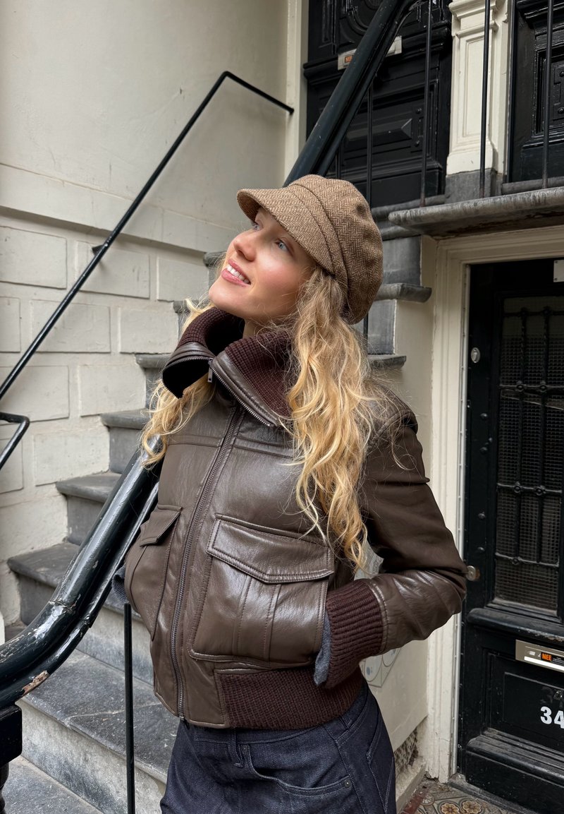 Chaqueta bomber de cuero marrón con puños de suéter acanalados, grandes bolsillos frontales y un cuello alto, acompañada de una gorra marrón texturizada.