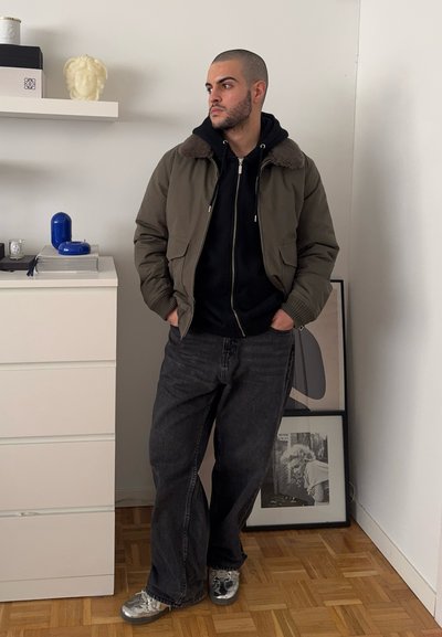 Chaqueta bomber verde oliva con collar de piel sobre una sudadera negra, combinada con vaqueros oscuros y zapatillas plateadas. Fondo liso y decoración mínima.