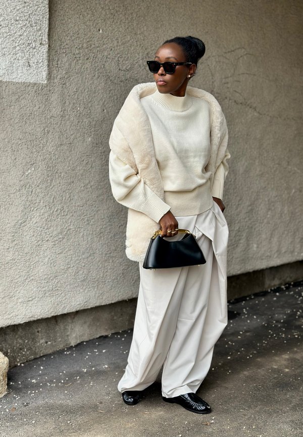 Nainen ylipitkässä valkoisessa neuleessa, leveissä kermaisissa housuissa, turkisviitassa, mustissa aurinkolaseissa ja mustassa käsilaukussa seisomassa teksturoidun seinän edessä.