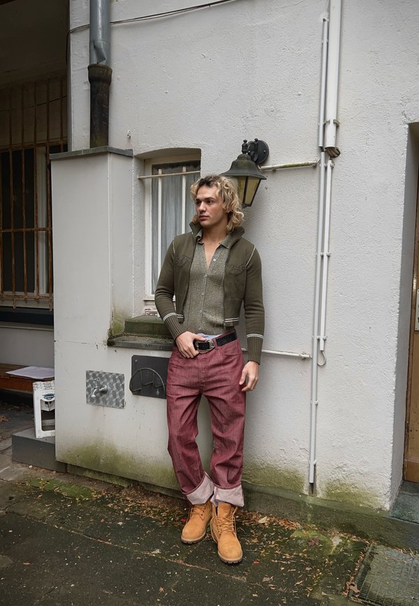 Uomo che indossa un cardigan lavorato a maglia verde oliva, un maglione chiaro e pantaloni rossi con i risvolti, abbinati a stivali da lavoro color sabbia, in piedi contro un muro.