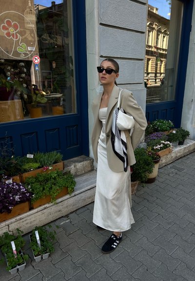 Blazer beige sobre un vestido de satén blanco, zapatillas negras, sosteniendo un bolso blanco con una bufanda estampada. Macetas y escaparate de la tienda al fondo.