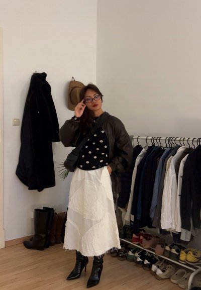 Mujer ajustándose las gafas, lleva una chaqueta oscura, blusa de lunares, falda blanca y botas negras, de pie junto a un perchero con ropa y abrigos, con zapatos en el suelo.