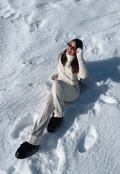 Conjunto atlético de color beige claro con chaqueta de cremallera y pantalones ajustados, sentado sobre nieve blanca y texturizada. Zapatillas negras con cordones y unas gafas de sol llamativas.
