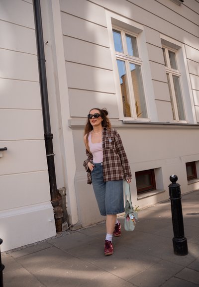 Mujer con gafas de sol, camisa de cuadros, top rosa y pantalones cortos vaqueros caminando por la acera cerca de un edificio beige con grandes ventanas.