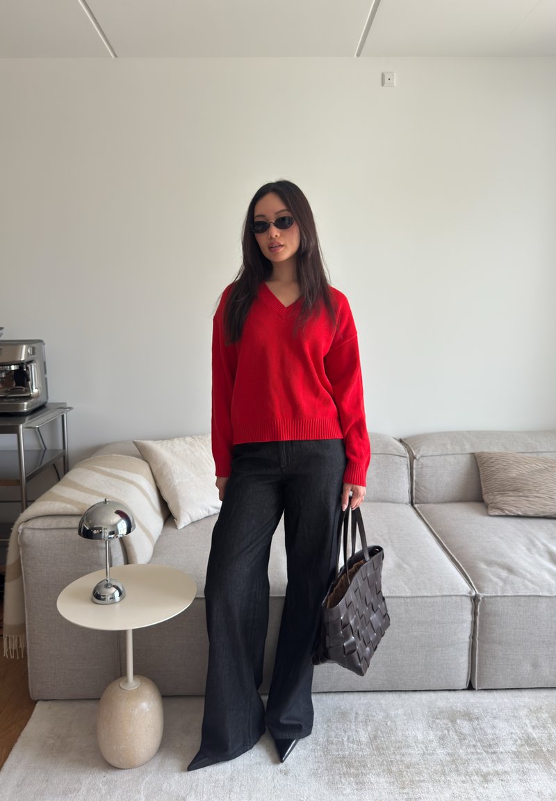 Femme aux cheveux longs portant un pull rouge, un pantalon noir large, des lunettes de soleil et un cabas en osier, debout près d'un canapé beige.