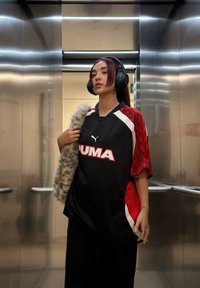 Černý sportovní dres s červenými a bílými akcenty, na kterém je velké logo "PUMA"; v kombinaci s černými kalhotami a šálou z umělé kožešiny; sluchátka přes hlavu.