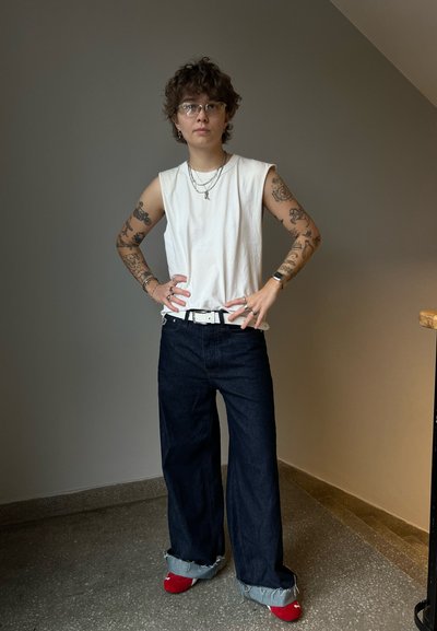 Camisa sin mangas blanca, pantalones vaqueros oscuros de pierna ancha con dobladillos deshilachados y zapatos rojos. Los accesorios incluyen collares en capas y anillos.