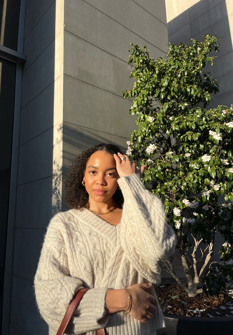 Pull à mailles torsadées beige avec un col en V, des manches longues et des poignets côtelés. Un sac à main marron est porté à l'épaule. Plante verte en arrière-plan.