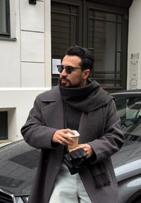 Sivi volneni plašč z visokim ovratnikom, oglen šal, črne rokavice in sončna očala, ki drži skodelico kave. V ozadju mokro vozilo in zid.