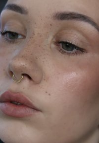Gros plan d'un visage à la peau claire, aux yeux verts, avec des taches de rousseur, un maquillage naturel et un piercing septum en forme de cœur doré.