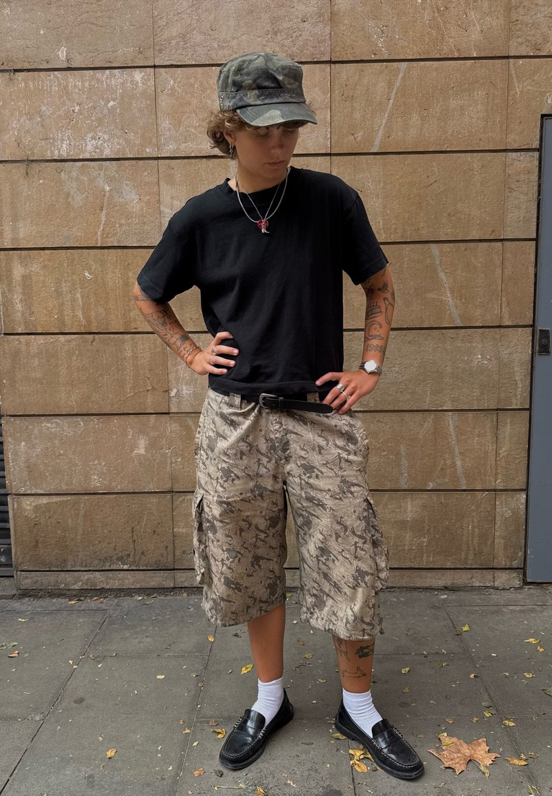 Pessoa com tatuagens vestindo um boné camuflado, uma t-shirt preta, calções de carga beige estampados, meias brancas e sapatos de couro pretos, em pé numa calçada.