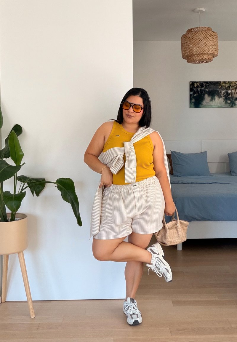 Kvinna i gul topp, randiga shorts och sneakers står på ett ben inomhus och håller en flätad handväska nära en krukväxt och säng.