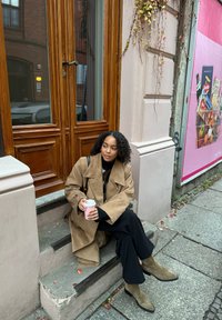 Beige semsket trenchcoat, svart turtleneck, sorte vide bukser, olivengrønne ankelstøvler; holder en lyserød kop, sittende på steintrapper.