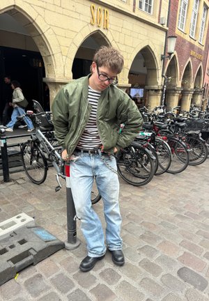 Chaqueta bomber verde sobre una camiseta de rayas blancas y negras, jeans claros desgastados y botas negras. El fondo muestra bicicletas aparcadas.