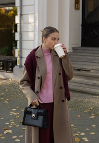 Casaco de lã castanho, suéter rosa, cachecol bordeaux e mala preta com acabamento fofinho. A segurá um copo de café branco, em p�é num passeio coberto de folhas.