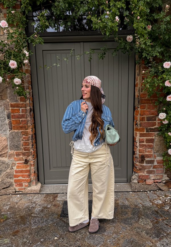 Jonge vrouw met hoofddoek, spijkerjack en wijde crèmekleurige broek staat glimlachend bij een grijze deur omlijst met baksteen en bloeiende roze rozen.