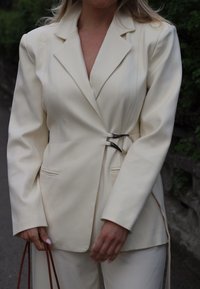 Blazer de couleur crème avec une coupe ajustée, présentant des revers nets et un design asymétrique, sécurisé par des accents en métal argenté.