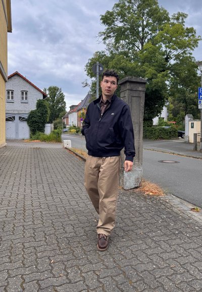 Un hombre camina por una calle pavimentada de piedras, vistiendo una chaqueta azul oscura, una camisa a cuadros, pantalones beige y zapatos marrones con cordones.