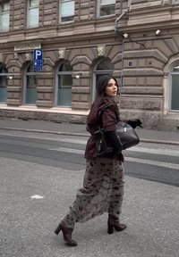 Giacca marrone aderente, guanti neri, gonna grigia con motivo floreale, stivali marroni con tacco e una grande borsa scura, mentre cammina lungo una strada della città.