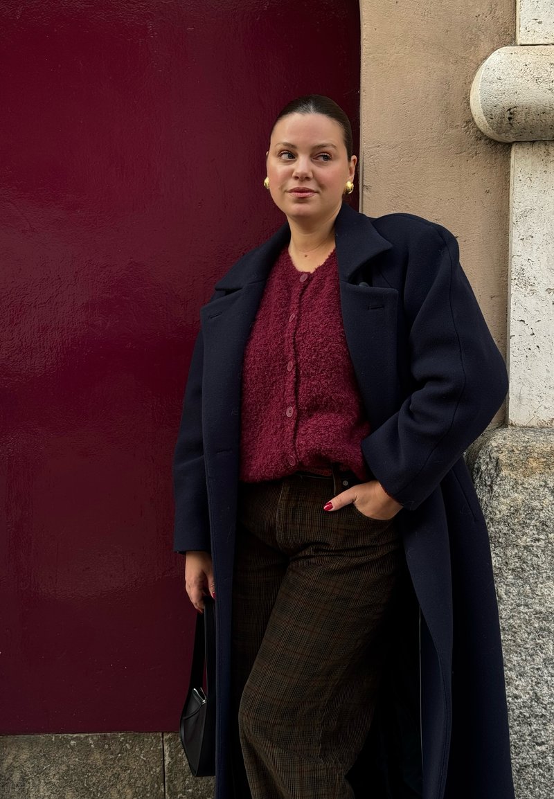 Manteau bleu marine surdimensionné, cardigan bordeaux en maille duveteuse avec boutons, pantalon marron à carreaux et petit sac à main noir. Boucles d'oreilles créoles en or.