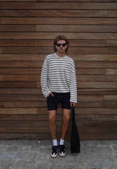 Suéter de rayas crema, pantalones cortos negros, bolso negro y gafas de sol oscuras. El calzado presenta un diseño en azul y blanco. Fondo de tablones de madera.