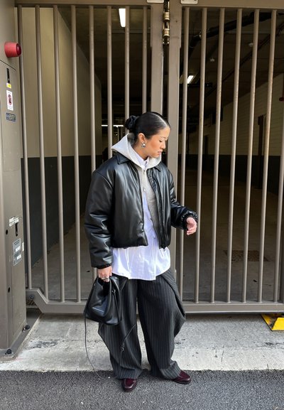 Mujer con chaqueta de cuero negra, camisa blanca, pantalones anchos de raya diplomática grises, sosteniendo un bolso negro, parada cerca de una puerta metálica alta en un entorno urbano.