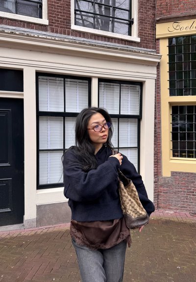 Joven mujer con cabello largo y negro, usando gafas, suéter negro y blusa marrón, llevando un bolso beige con patrón, en una calle pavimentada con ladrillos.