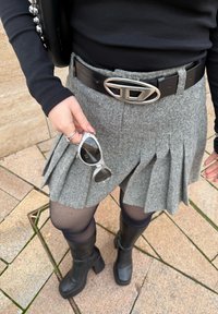 Grå plisserad kjol med ett svart läderbälte med ett silverfärgat spänne, kombinerat med svarta knähöga stövlar och håller i solglasögon.