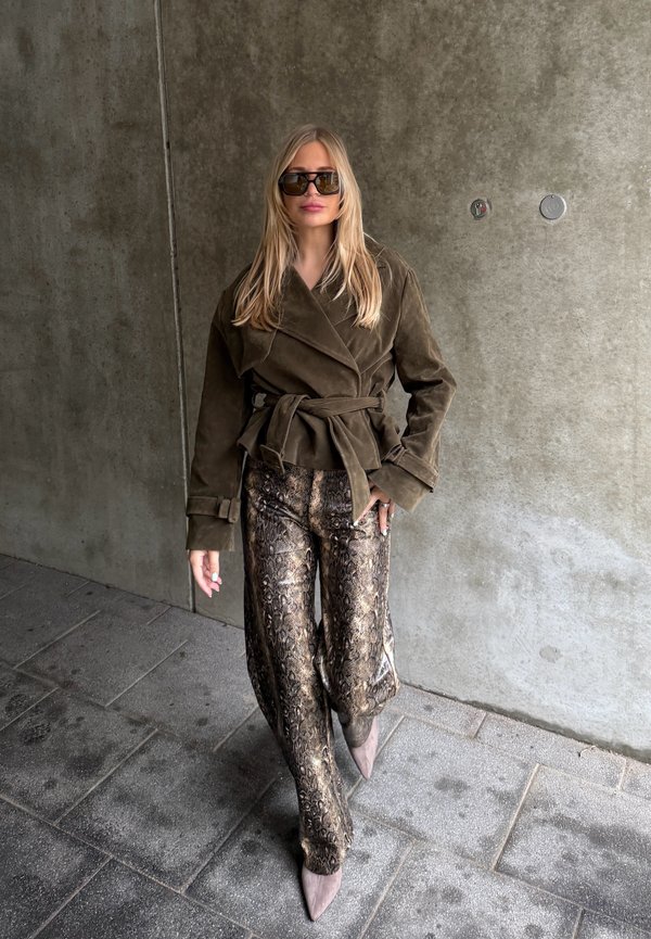 Donna con capelli biondi lunghi che indossa occhiali da sole oversize, una giacca marrone con cintura, pantaloni con stampa a serpente e scarpe beige con punta, in piedi sul marciapiede.