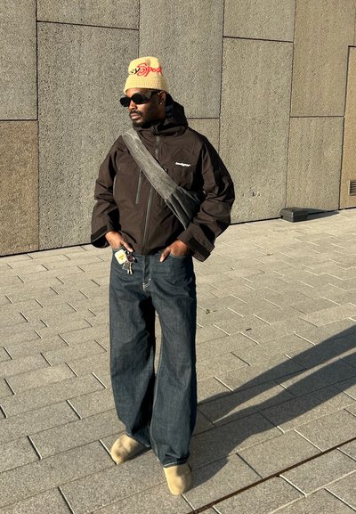 Chaqueta negra impermeable con bolsillos con cremallera, pantalones vaqueros de mezclilla azul de corte holgado, sombrero beige con logo y zapatos de lona color tan claro.