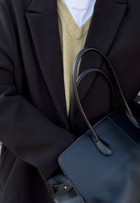 Borsa a mano in pelle nera con texture liscia, doppi manici e forma strutturata, tenuta contro un cappotto scuro e un maglione beige chiaro.