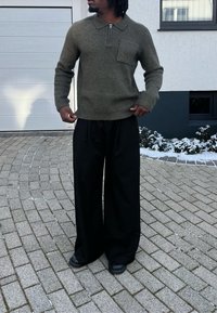 Personne portant un pull à manches longues vert olive et un pantalon noir à jambes larges, debout sur une allée pavée près d'un parterre de jardin saupoudré de neige.