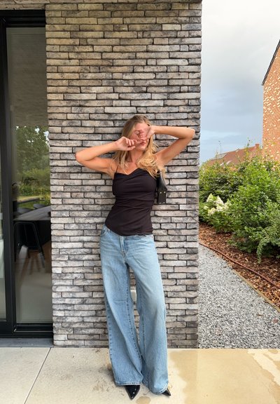 Top negro sin mangas con diseño en capas, combinado con jeans de pierna ancha en azul claro. Tacones de punta negra y un pequeño bolso negro. Fondo de pared de ladrillo.