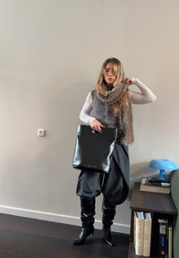 Femme portant une écharpe en fourrure, un haut blanc à manches longues, un gilet gris, un pantalon noir à fines rayures, et des bottes noires, tenant un sac fourre-tout en cuir noir à l'intérieur.