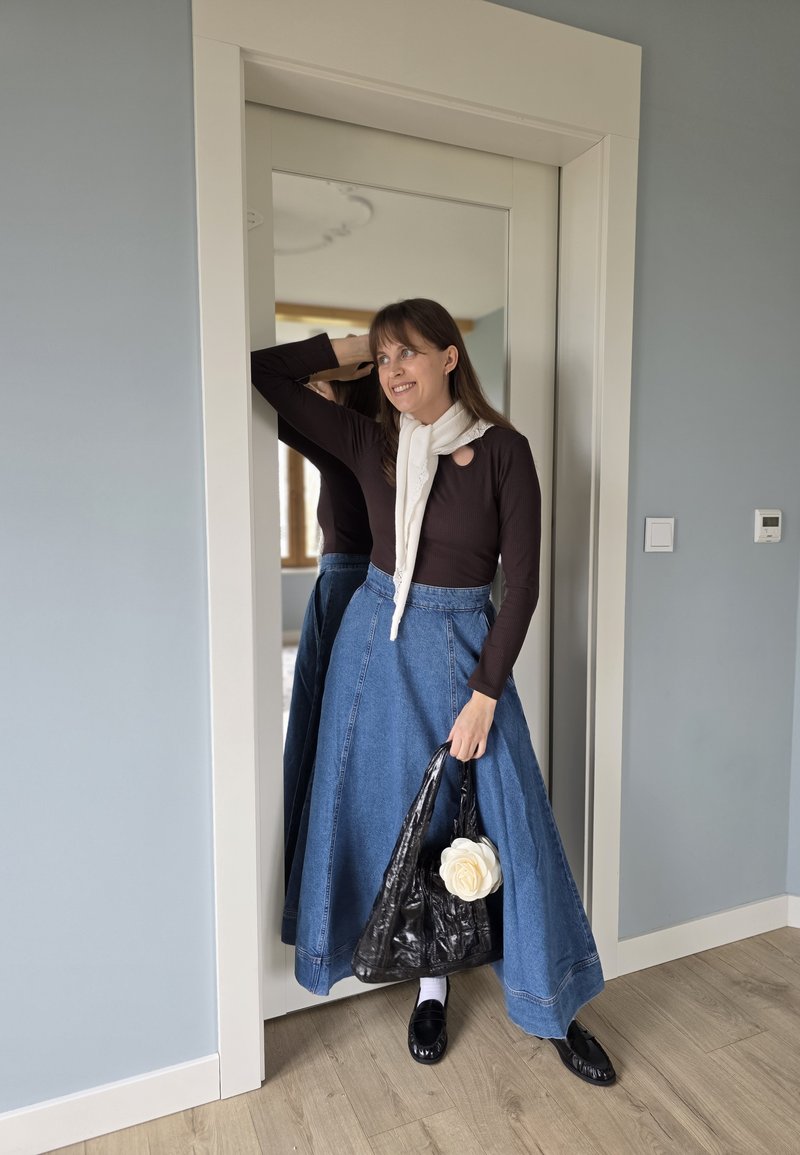 Femme en jupe longue en jean et haut sombre se tient près d'un miroir, tenant un sac noir avec une fleur blanche, souriante et reposant son bras sur la tête.
